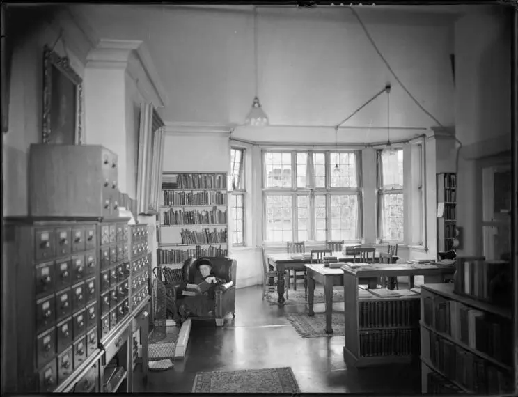 Black and white photo shows a reading room and a view of drawers of catalogue cards, a bookcase, and tables and chairs, towards a bay window. An elderly woman sits reading a book in a chair by a fireplace.