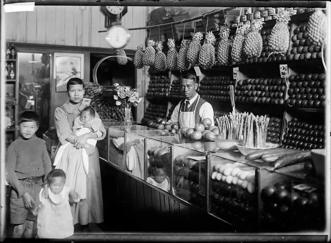 Shopkeeper, Wong Gar Sui, stands behind a counter with asparagus, apples and cucumbers on top. Above their head hang a row of pineapples. Wong Gar Sui’s wife, Mew Yuen, stands with their 3 children.