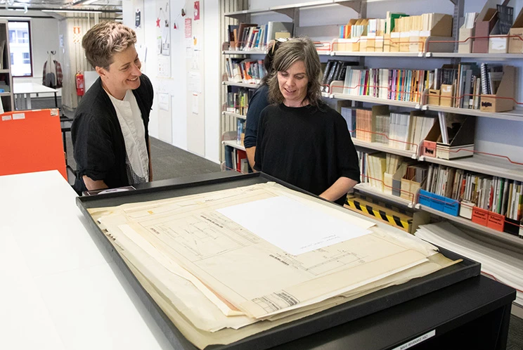 Two people in a room with large bookshelves standing at a work table looking at some papers.