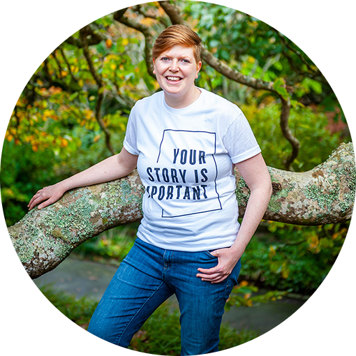 A woman wearing jeans and a white t-shirt with the words 'Your story is important', leaning against a tree branch.