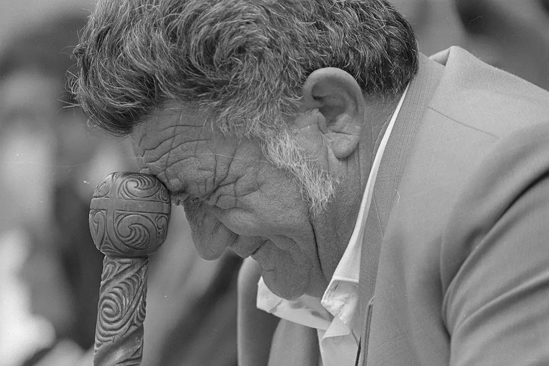 Black and white, close-up photo of Mr Mac Taylor, Ngāpuhi elder, bowing his head and leaning on his walking stick.