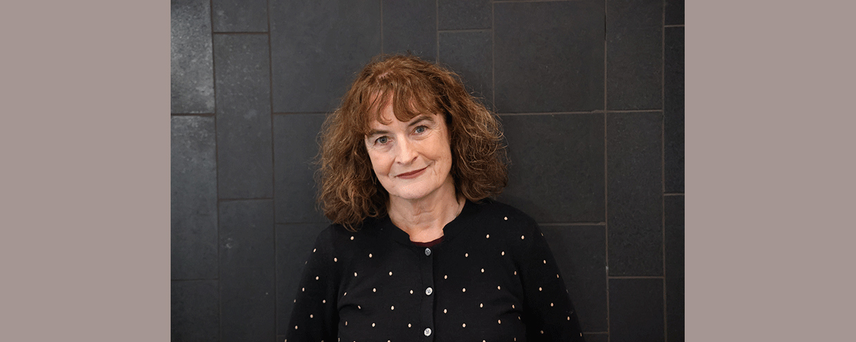 Photo of Kate De Goldi wearing a polka-dot cardigan, standing against a dark tiled wall.
