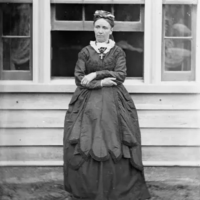 Victorian era black and white photo a woman standing outside a weatherboard house. Her arms are crossed she is looking at the ground. 