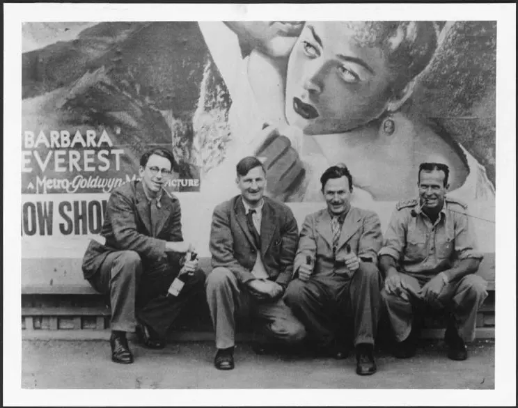 Black and white photo of a group of four men, crouching against a wall for a group portrait. 