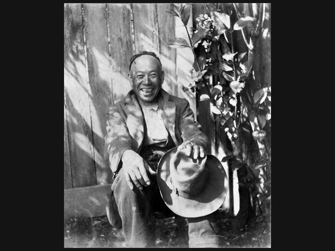 Black and white photo of Joe Tie sitting next to a fence and smiling at the camera.