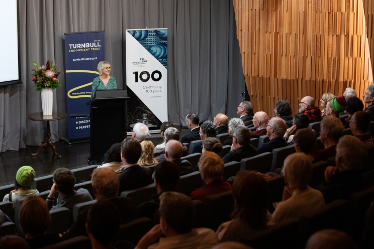 A woman stands behind a lectern delivering her speech while the audience looks on from their seats.