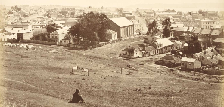 A man seated on a hill overlooking Auckland city with wooden buildings and dirt streets.