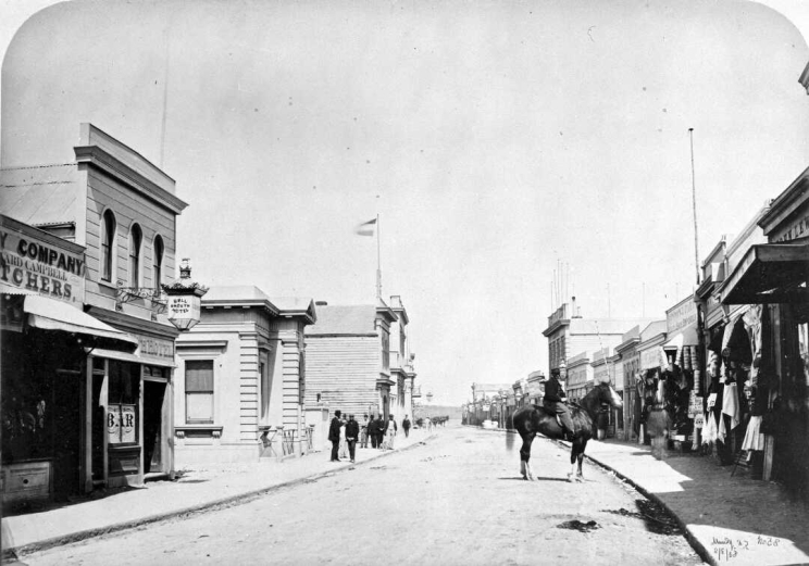 On a street lined with wooden buildings a horse and rider stand sideways to the camera, plus there are a number of pedestrians visible on the footpaths.