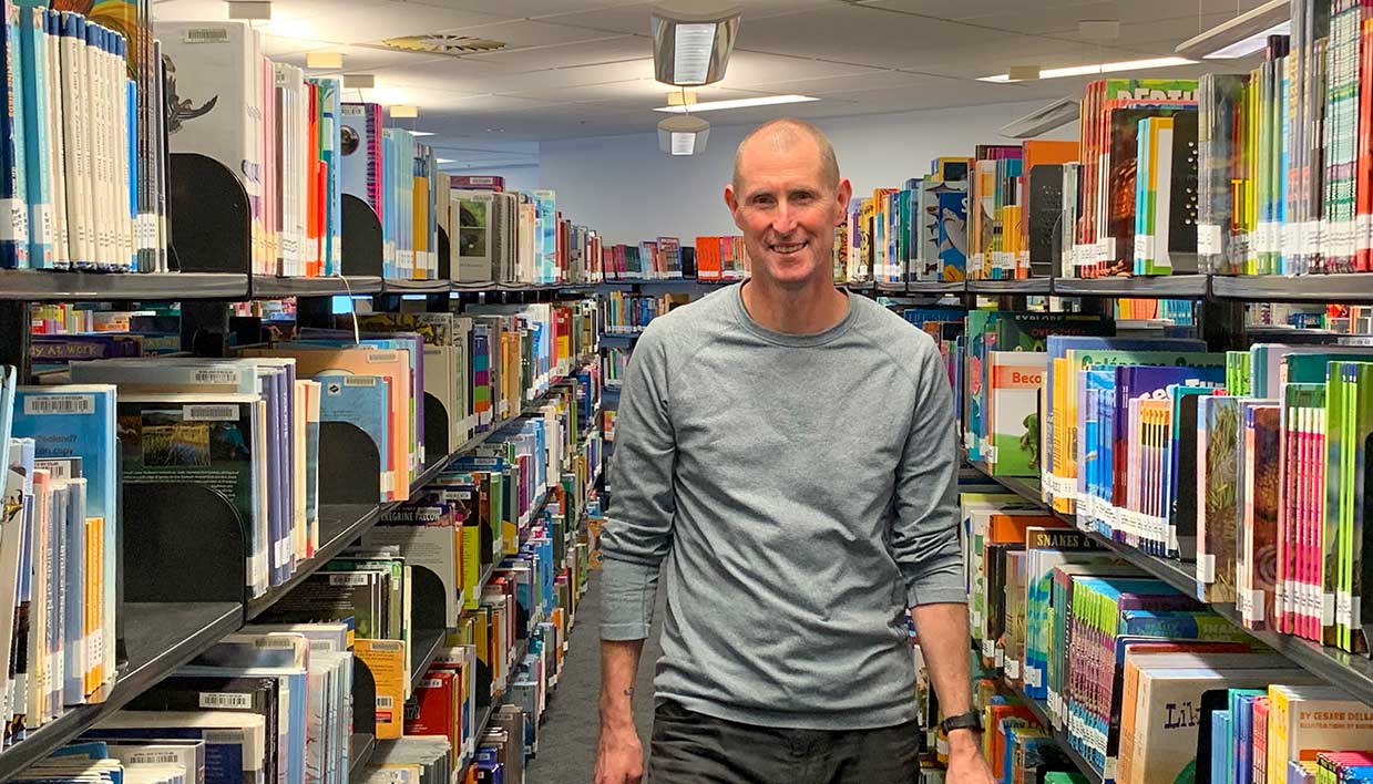 Alan Dingley holding a book and walking between shelving at the National Library Auckland centre.