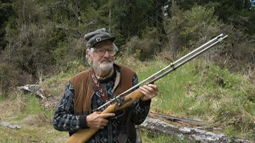 Bearded man wearing a hat holding a shotgun.