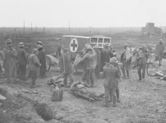 General view of an advanced dressing station on battleground at 'Somme Farm', Ypres Salient. Scene includes soldiers gathered around an ambulance and a wounded soldier laying on a stretcher on the muddy ground.