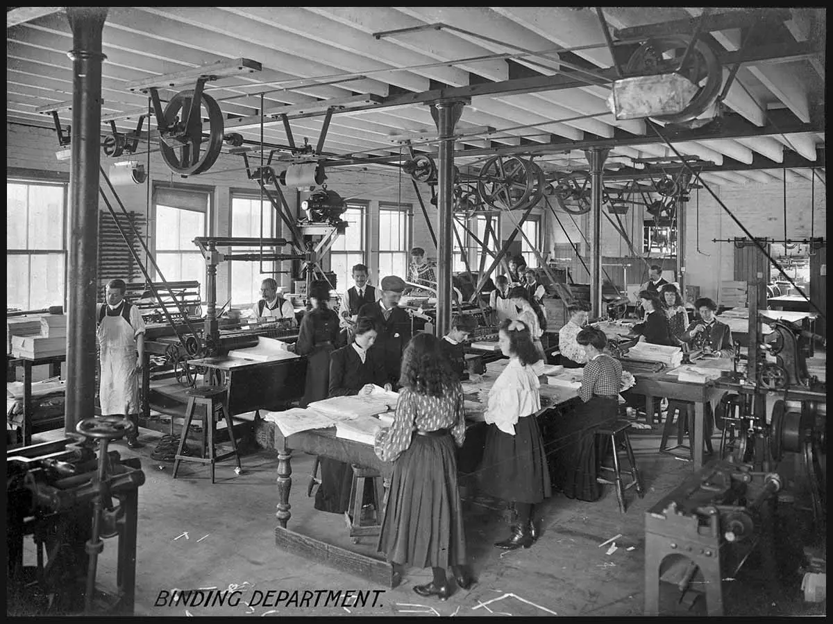 Workers are standing or sitting at tables looking at papers in the binding department of ‘The New Zealand Herald’ newspaper. Machinery is on some of the tables and is also hanging from the ceiling.