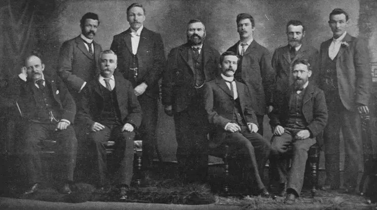 A formal portrait of a group of ten men, some seated and some standing. 