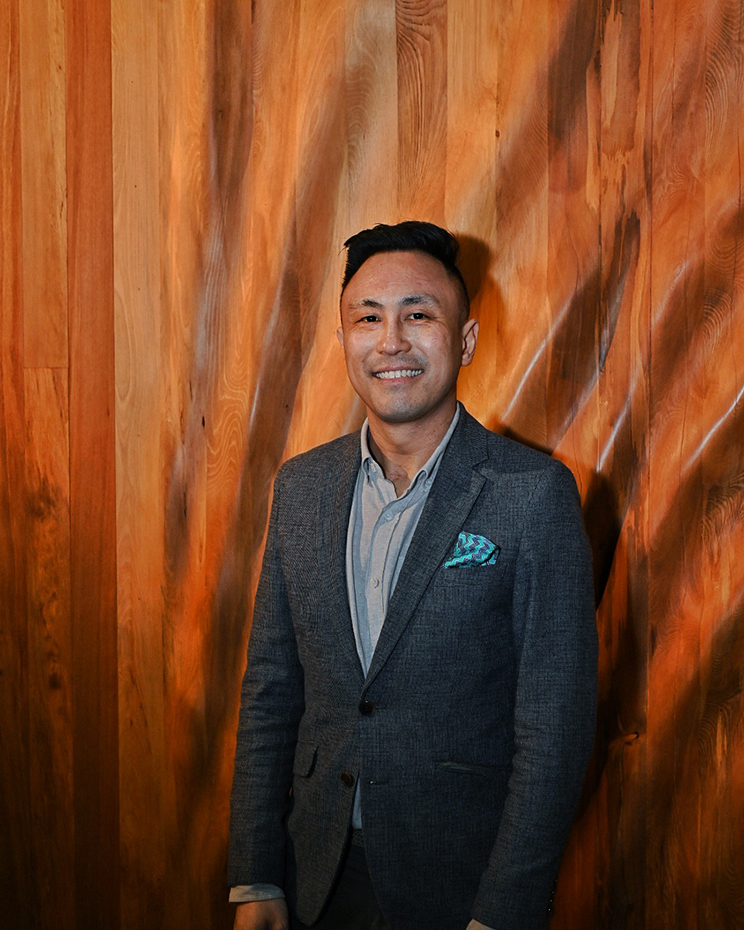 Smiling Asian man standing in front of a curvaceous wooden wall.