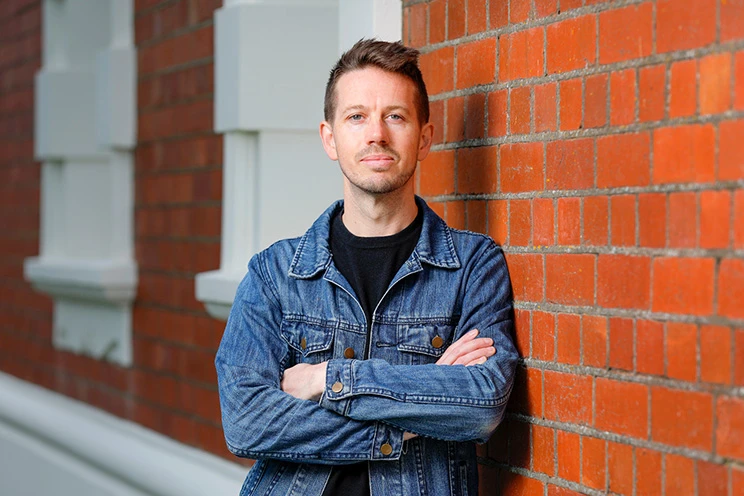 A man wearing a blue denim jacket and black t-shirt leaning on a brick wall.