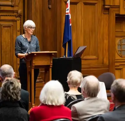 Dr Lydia Wevers standing at a podium in the Great Hall at Parliament. 