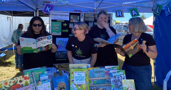 4 people in black t-shirts display colourful children's books at an outdoor literacy promotion stall under a blue marquee.