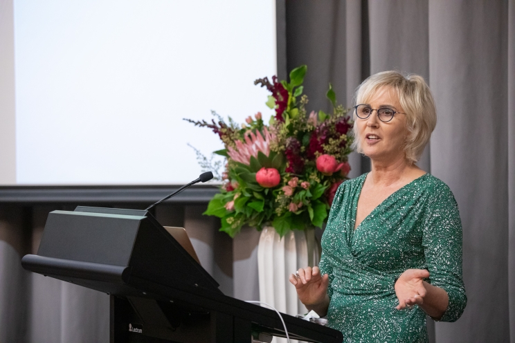 A woman in a green dress stands behind a lectern delivering her speech while gently gesticulating with her hands.