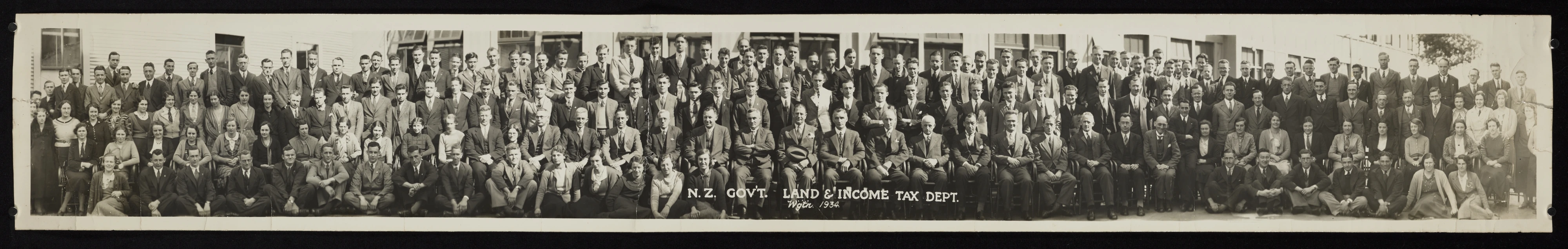 Panoramic group portrait of over 100 employees of the Land and Income Tax Dept., they are positioned about 4-5 rows deep, some in front row are seated on ground.