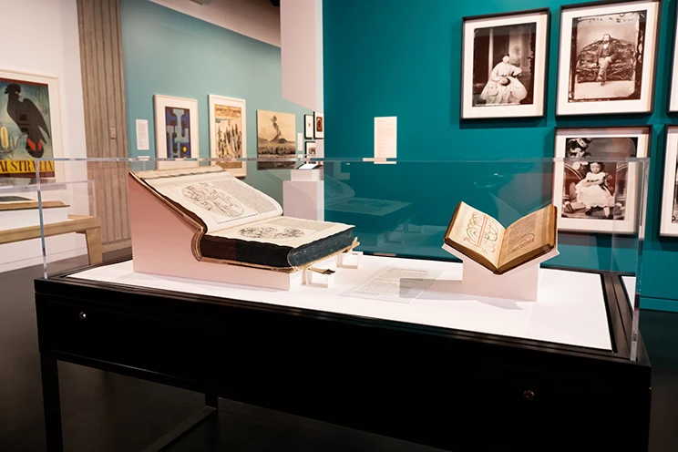 Two open books on display in a glass cabinet with a number of photographs and posters behind on the walls in an exhibition.