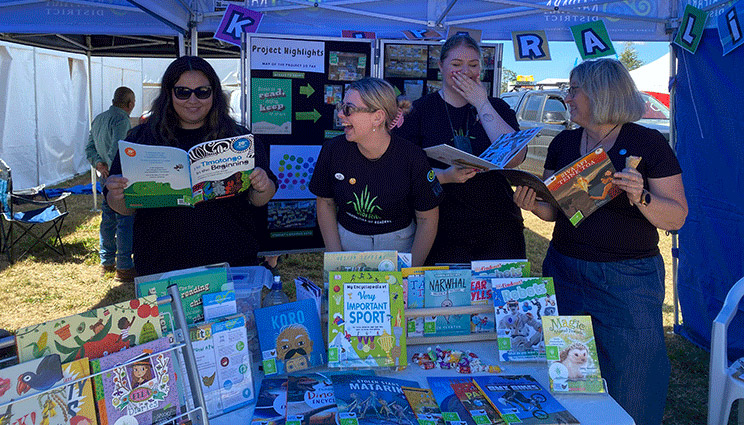 4 people in black t-shirts display colourful children's books at an outdoor literacy promotion stall under a blue marquee.