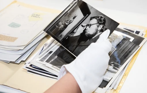 A person wearing white gloves holds a black and white photograph from a folder containing a stack of photos.