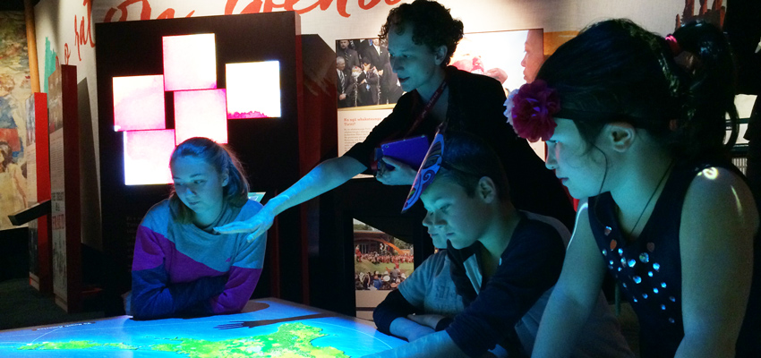 School children with a Learning Facilitator gathered around a display table at the He Tohu exhibition in Wellington