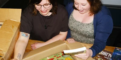 School loan coordinator and teacher smiling as they look at some National Library school lending service books they're unpacking from a box. 