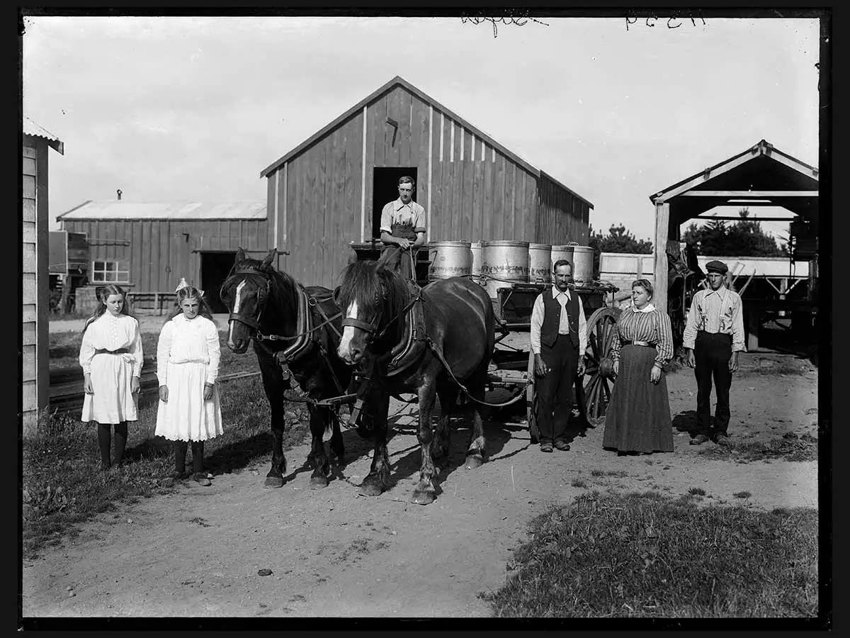 Members of the Styles family standing on their dairy farm. A person is sitting on a cart carrying large cans of milk, holding the reins of 2 horses that are pulling it. In the background are large farm sheds.