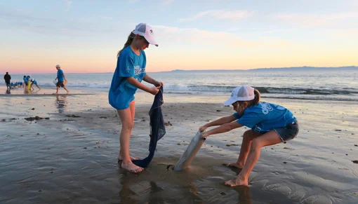 Two primary school students on a beach participating in a science project using ocean research equipment on the sand at sunset with water and other figures in the background.
