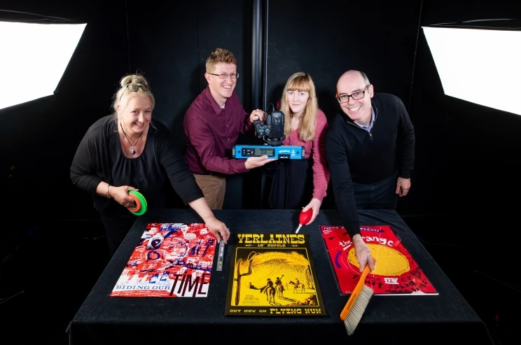 A team photo taken in the studio with four technicians showing three colourful posters along with the camera gear and stand they use for the photography.