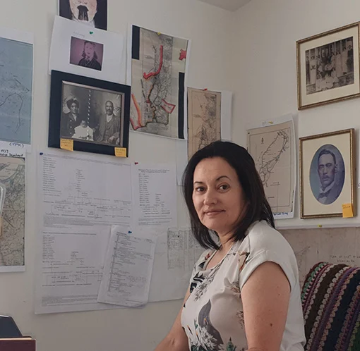 Portrait of a woman sitting in a room with maps, notes and old photographs on the wall behind her.