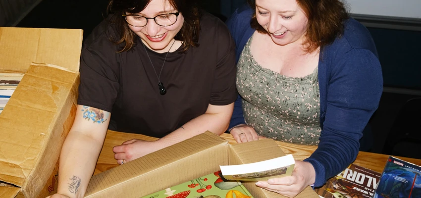 School loan coordinator and teacher smiling as they look at some National Library school lending service books they're unpacking from a box.