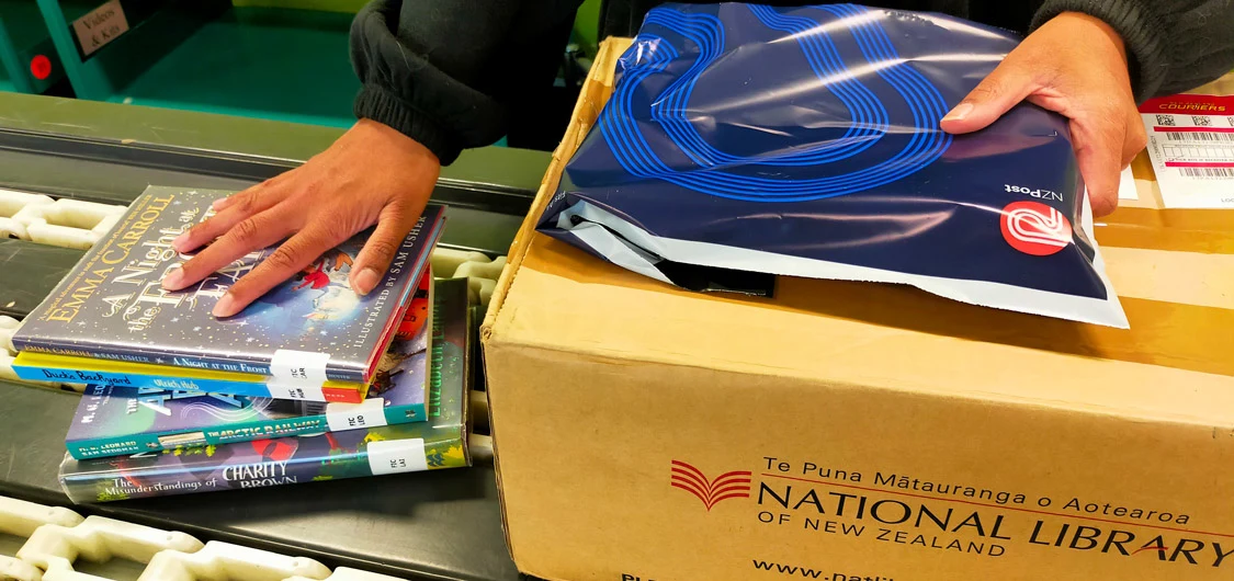 Processing National Library's lending service book returns. Shows a woman's hands, and also shows books, a courier bag and a National Library box on a conveyor belt. 