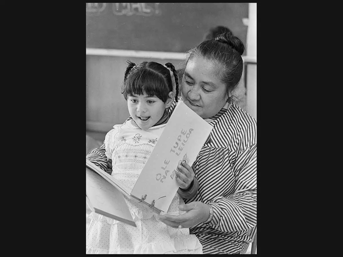 Fereni Ete reading a story in Samoan to a child, Fuatina Mapu. In the background is a blackboard on the wall with chalk writing on it.