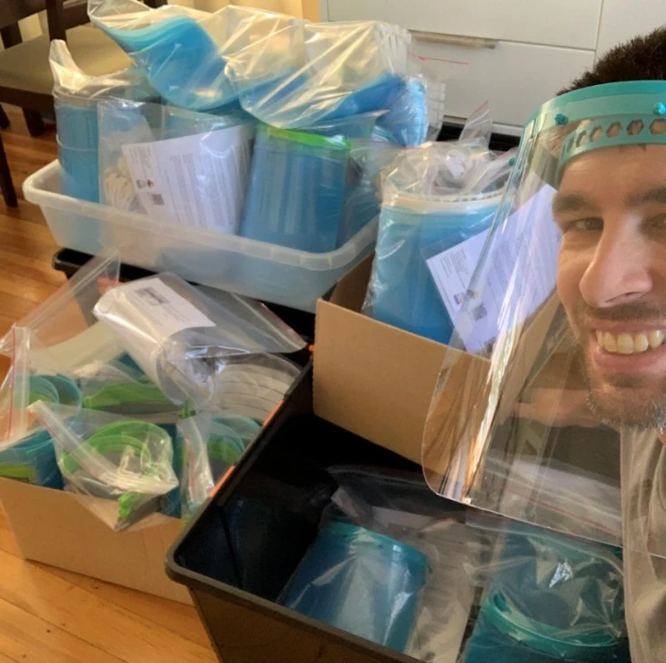 A man wearing a face shield and smiling with hundreds of similar face shields in the background, packaged and ready to distribute.