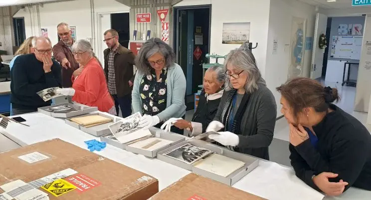 A group of men and women are assessing black and white prints held in archival boxes that are arranged on a large table. 