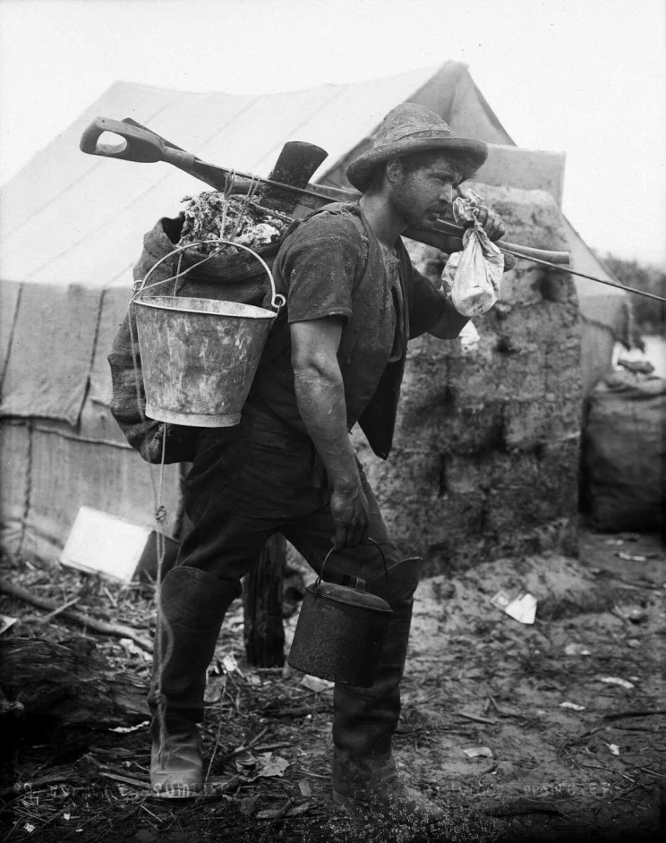 A man stands while supporting a heavy load with a bucket and large pīkau (a backpack made from a grain sack).