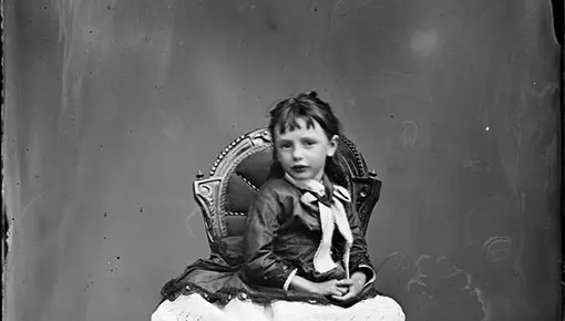 Victorian era back and white photo of a young girl sitting on a chair for a studio portrait. 