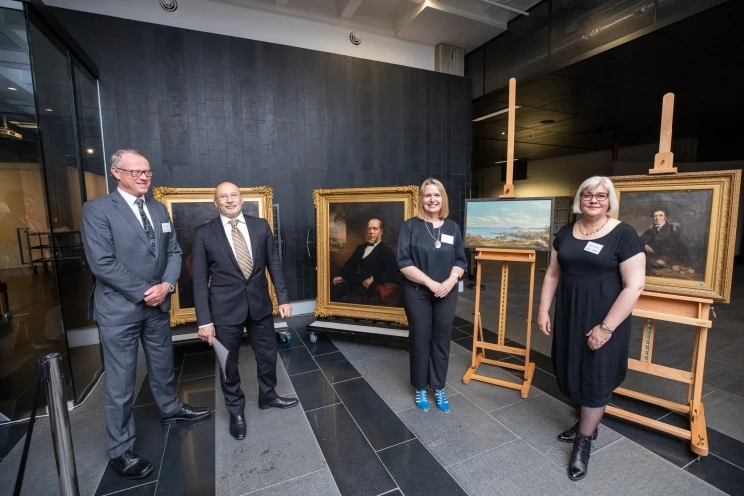 Four people in formal dress pose in front of four framed oil paintings.