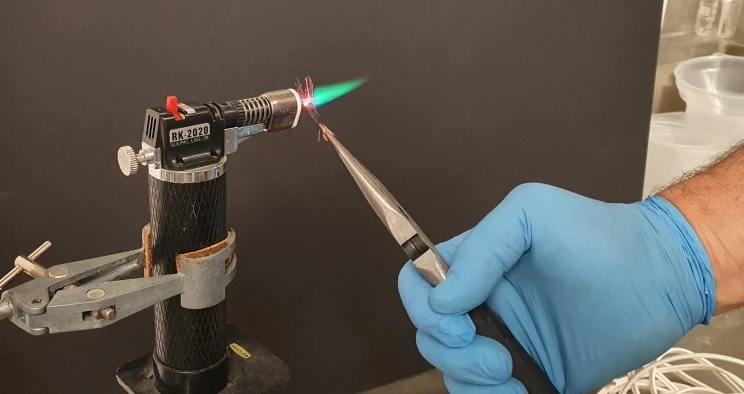 Someone wearing a rubber glove holds a fragment of copper wire in a flame using needle-nose pliers — the flame is turning green-coloured from a reaction with the wire.