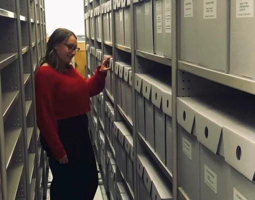 Shows the author in the stacks with archival boxes labelled neatly and in rows along shelves. 