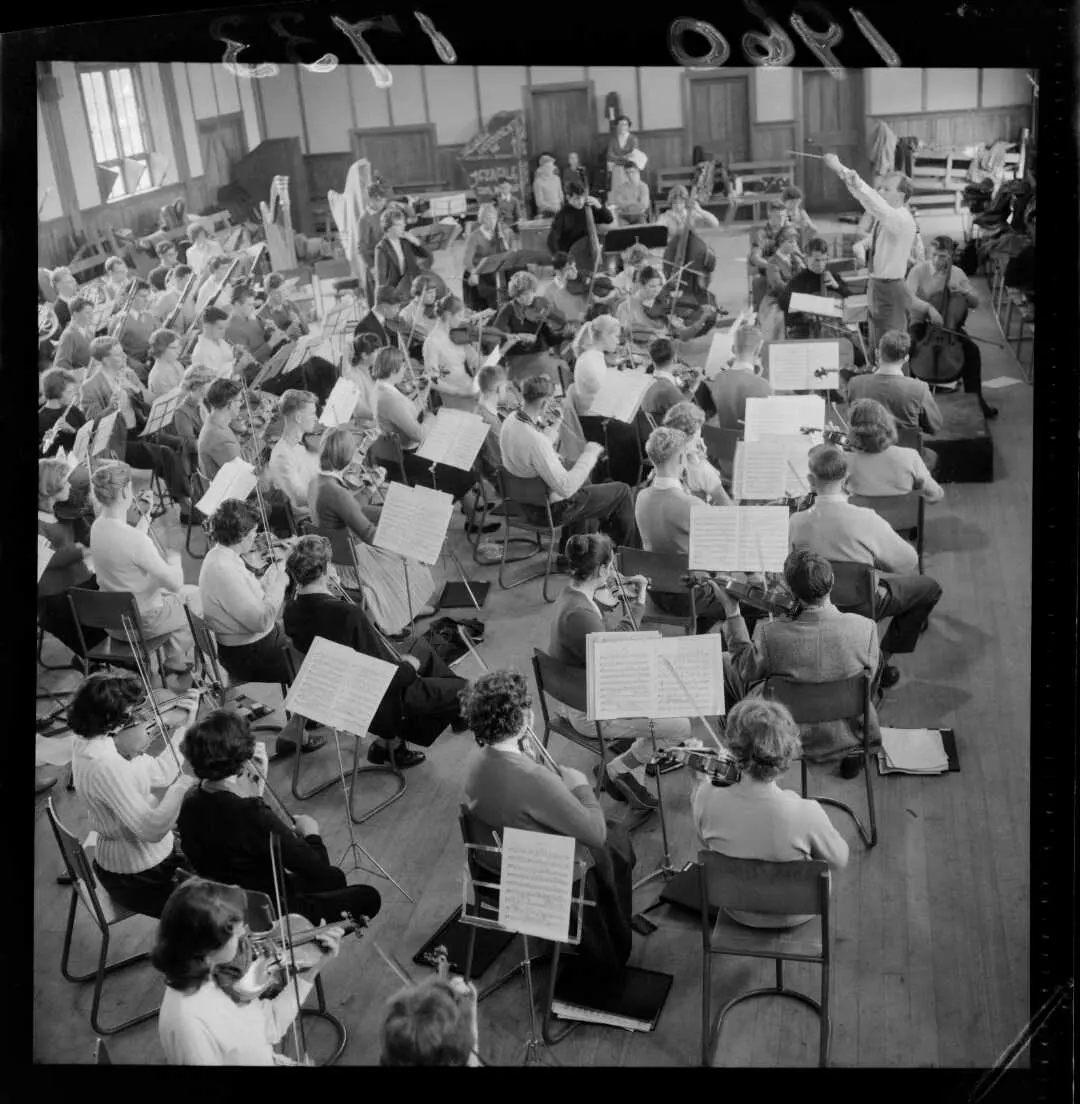Black and white photo taken from the back of a group of people playing musical instruments with a conductor at the front. 