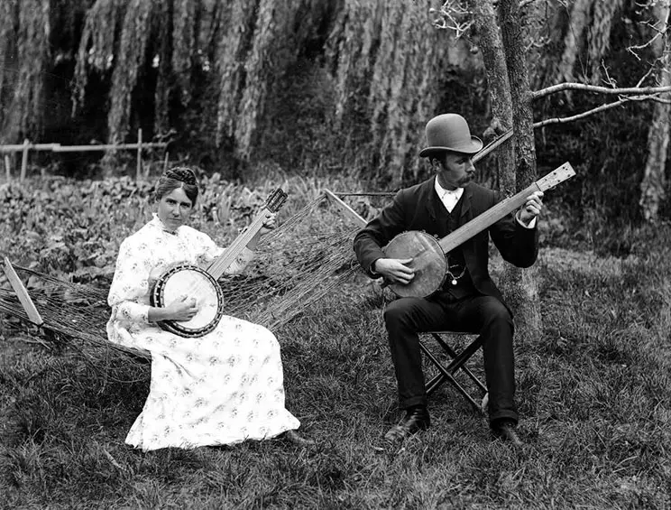 Black and white photo from Victorian-era showing a man and woman playing banjos outside. The woman is sitting on a hammock. 