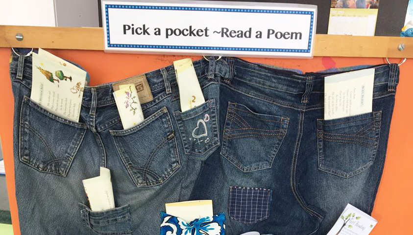 A sign 'Pick a pocket — Read a Poem' with a display of jeans hanging on a wall with poetry sticking out of the pockets