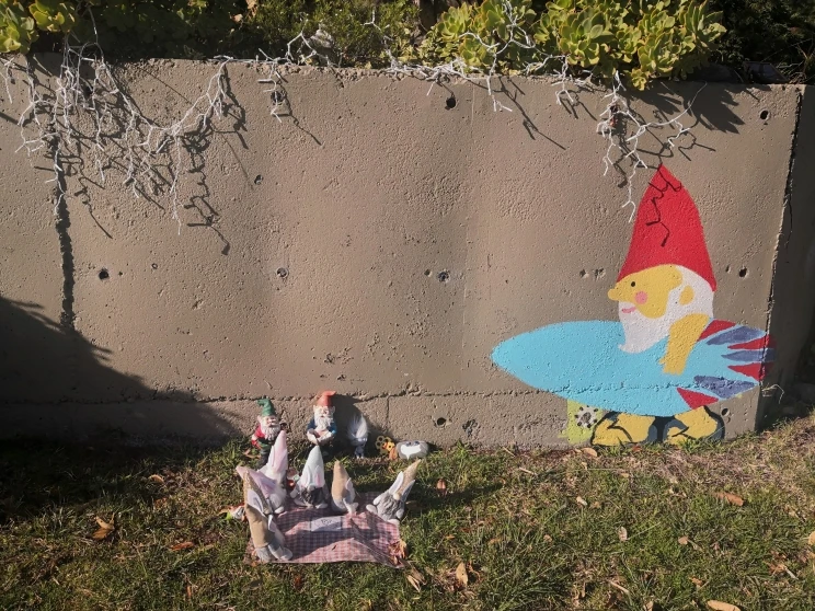 Gnome painted on concrete wall holding a surfboard and a gathering on the grass in front of the wall of small figurines appearing to have a picnic.