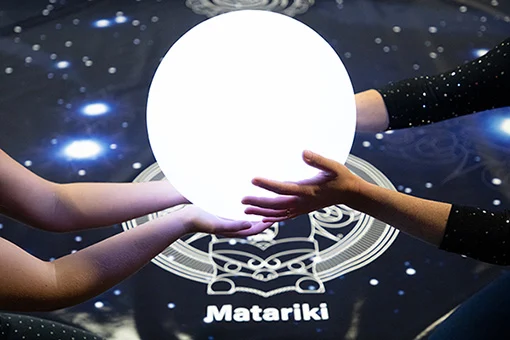 A glowing globe being held over a floor with stars and the word 'Matariki'.