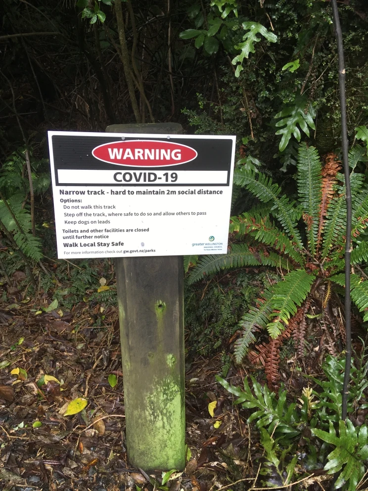 DOC signage for narrow tracks in parks and forests displayed on a white sign — "Warning COVID-19: narrow track - hard to maintain 2m social distance".