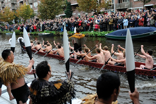 Nine Māori men paddling a waka taua (ceremonial canoe) down a river.