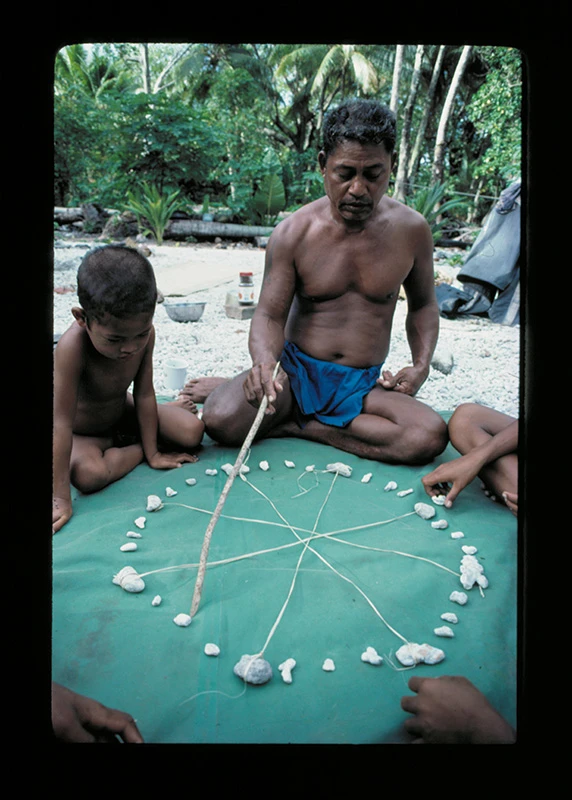 Colour photograph of Mau Piailug with his son, teaching navigation to a group, using a star compass made of shells and string.